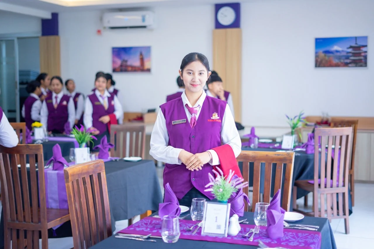 Mahasiswi The HIS berdiri di samping meja restoran dengan gelas dan serbet ungu.